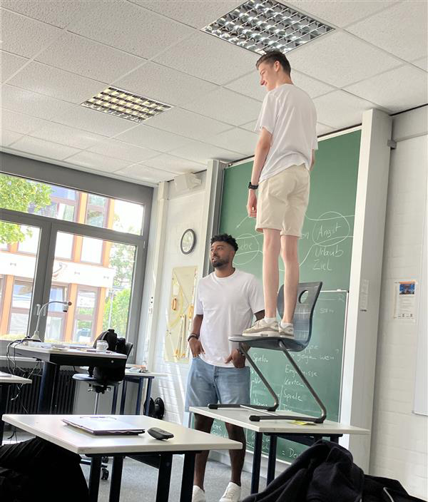 Ein Schüler steht auf einem Stuhl, welcher auf einem Tisch steht, im Klassenzimmer mit Trainer