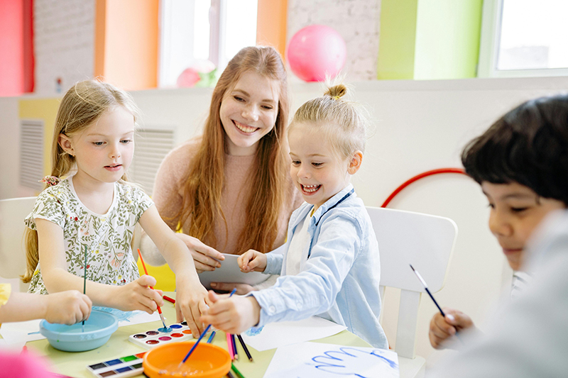 Erzieherin mit Kindern an einem Tisch. Kinder malen mit Farben