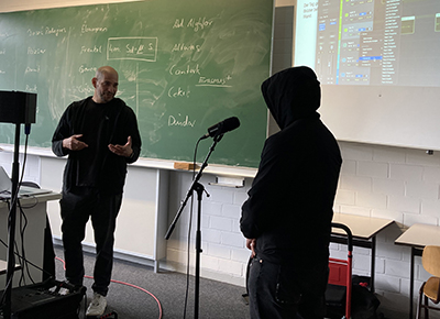 Bild eines Klassenzimmer, Rapper vor einer Tafel und Schüler am Mikrophon