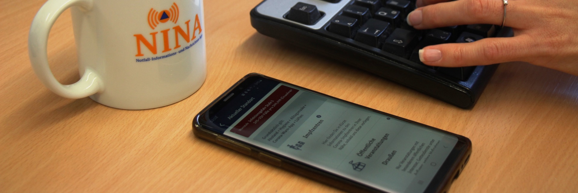 Auf einem Tisch steht eine Tasse, daneben liegt ein Smartphone auf dem die Warn-App NINA geöffnet ist