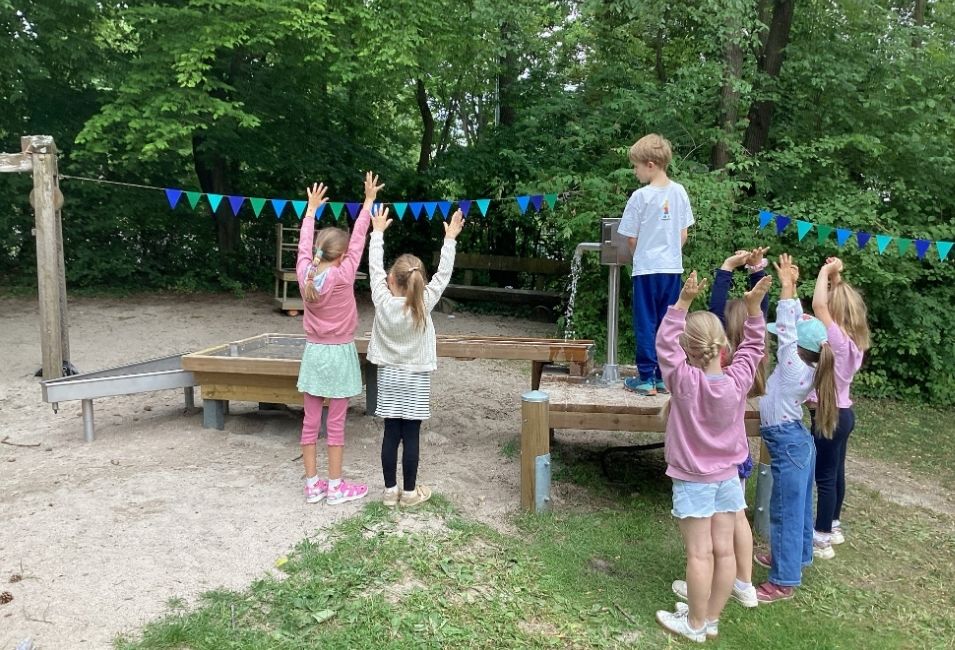 Ein Junge steht am Wasserhahn des Wasserspielplatzes. Andere Kinder stehen darum herum und heben die Hände in die Luft.