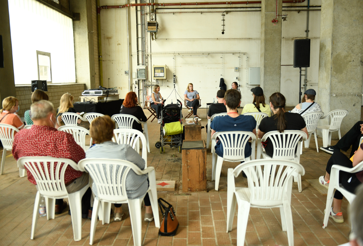 Man sieht eine Gruppe Menschen auf weißen Stühlen sitzen, die einem Gespräch lauschen. 