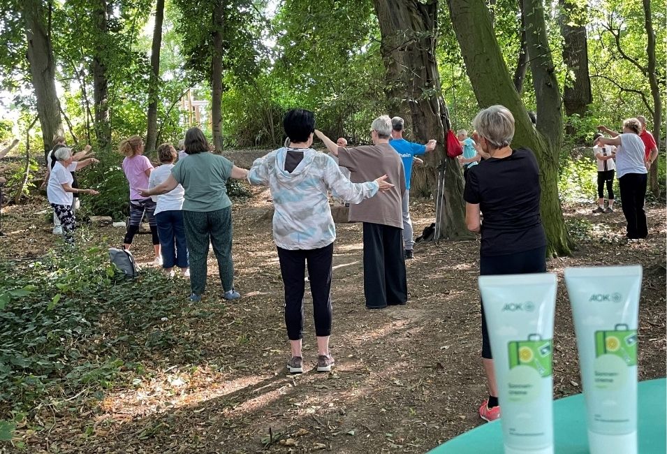 Eine Gruppe von Personen praktiziert Qigong im Schatten eines lichten Waldes. Im Vordergrund liegen zwei weiße Sonnencremetuben mit grünen Etiketten und der Aufschrift AOK.