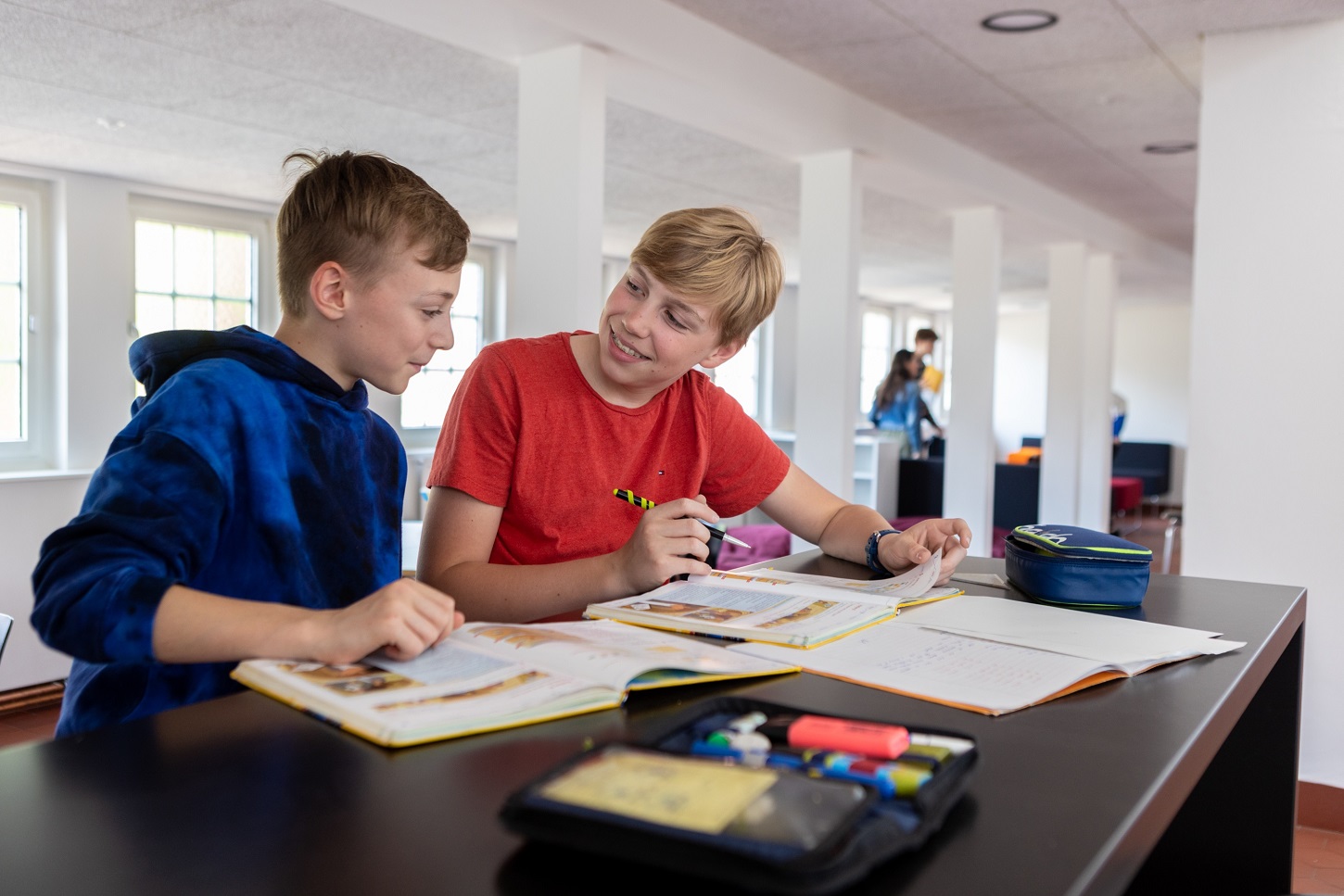 Zwei Schüler sitzen an einem Tisch und arbeiten an Hausaufgaben oder lernen. Vor ihnen liegen Bücher und Notizbücher.