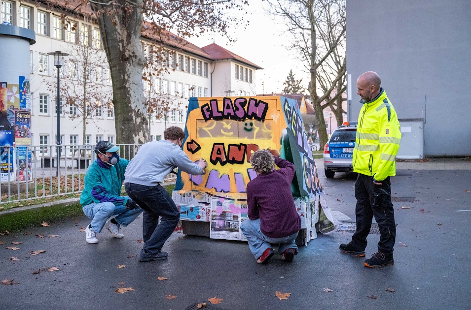 Drei Jugendliche besprühen einen Blitzeranhänger. Rechts daneben steht ein Mann mit einer gelben Warnjacke.