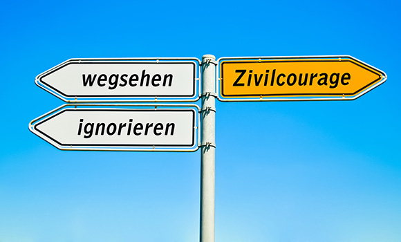Drei Wegweiser an einem Mast. Einer hat die Aufschrift "Zivilcourage". Die anderen beiden Wegweiser zeigen in die entgegengesetzte Richtung und tragen die Aufschrift "wegsehen" beziehungsweise "ignorieren".