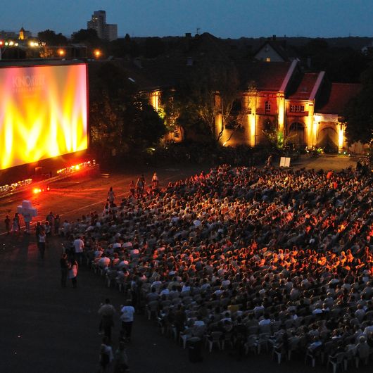 Viele Menschen sitzen auf Stühlen vor einer Kinoleinwand, die draußen steht.