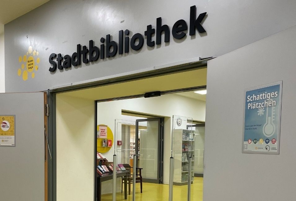 Eingangsbereich der Stadtbibliothek. An der geöffneten rechten Türe hängt ein Plakat mit der Aufschrift „Schattiges Plätzchen“.