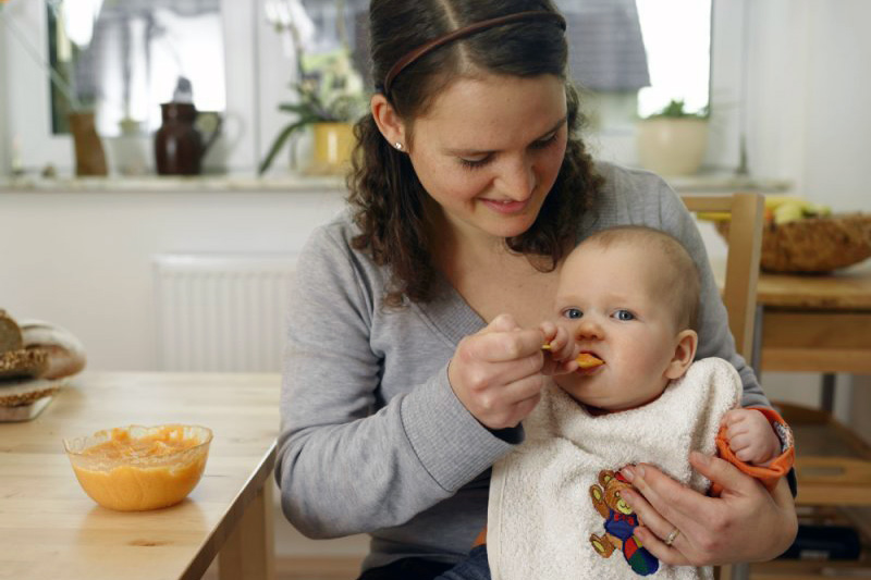 Mutter mit S&auml;ugling auf dem Scho&szlig; f&uuml;ttert Brei