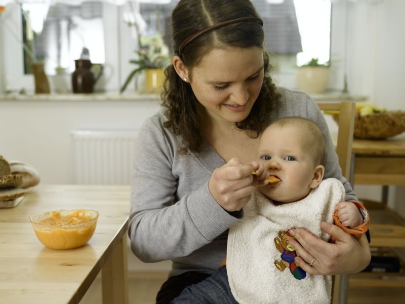 Baby sitzt auf dem Schoss, Mutter f&uuml;ttert es mit L&ouml;ffel 