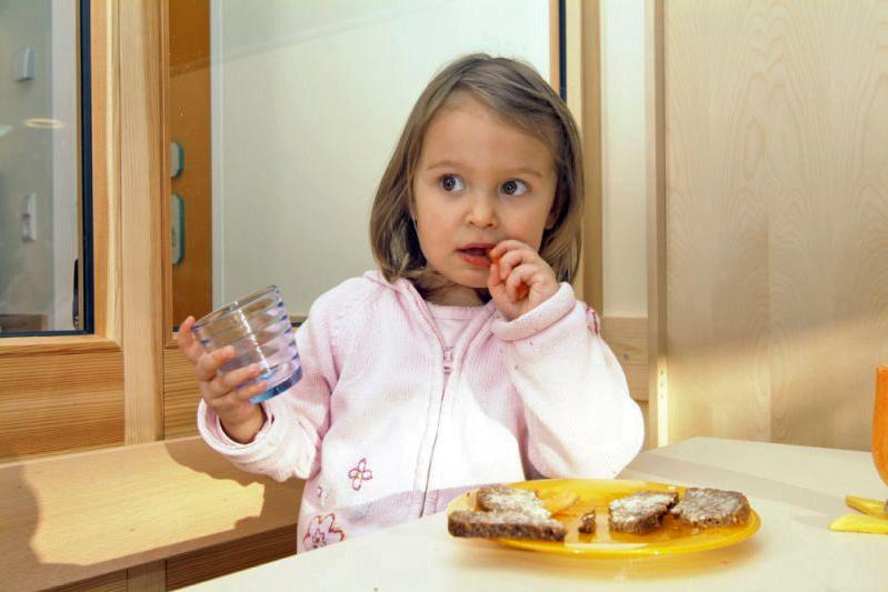 Kind in rosa Jacke mit Glas in der Hand, auf dem Tisch ein Teller mit Brot  